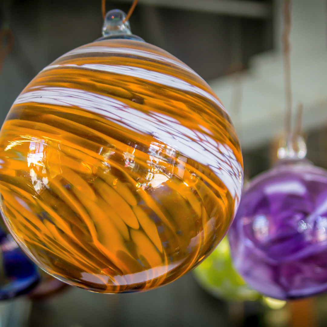 image of orange circular blown glass ornament in the foreground and a purple circular blown glass ornament in the back ground