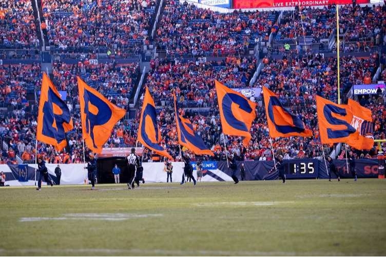 people running on Broncos football field holding large flags that spell out B-R-O-N-C-O-S stadium full of fans in background